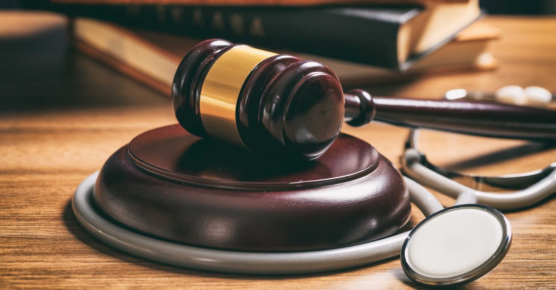 A judge's gavel rests on a wooden block next to a stethoscope, with law books in the background on a wooden surface.