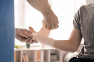 Hands of clinician in uniform wrapping right palm and wrist of sick patient with flexible bandage during medical procedure in modern clinics