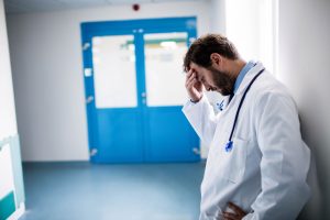 Male doctor leaning against pale wall pressing forehead in hospital corridor near red alarm bell. Healthcare, medical, stress, exhaustion, hospital, professionalism, clinical