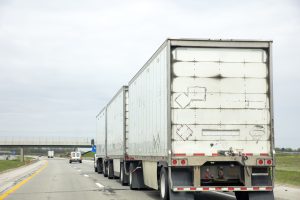 Three trailer road train transporting commercial cargo in USA highway. White long heavy vehicle rear, travels with cloudy sky background.